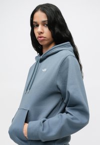 Γκρίζα-μπλε φούτερ με κουκούλα, μπροστινή τσέπη, και μικρό λογότυπο στο στήθος. Μαλακό ύφασμα με άνετη εφαρμογή και ριμπ ύφανση στους καρπούς.