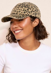 Gorra de béisbol con estampado de leopardo fabricada en tela, que presenta un visor curvado y detalles de costura, con una gama de colores en tonos neutros de marrón claro y negro.