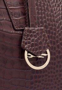 Donkerbruin, gestructureerd krokodillenpatroonleer, met een gouden embleemtag met de tekst "Paul Costelloe" en decoratieve stiksel.