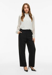 Lys, gennemsigtig hvid bluse med høj hals og elastiske manchetter, parret med skræddersyede sorte bukser og sorte højhælede sko med detaljer.