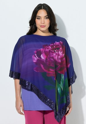 Frau trägt einen transparenten blauen Poncho mit großem pinkem Blumenmuster und Paillettenrändern über einem lila Top und rosa Hose.