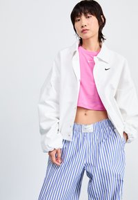 Bijela oversized jakna, ružičasti kratki majica, i plave široke hlače s elastičnim pojasom i Nike logom na majici i hlačama.