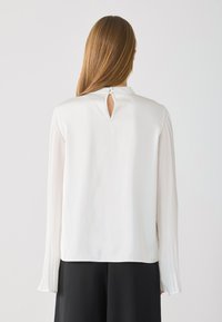 White blouse with pleated long sleeves, keyhole back detail, and a smooth texture. Complemented by a minimalistic design.