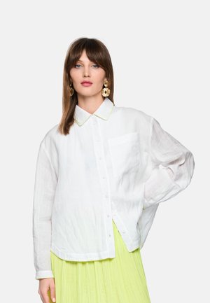 Vrouw met stijl bruin haar draagt een oversized witte blouse met knopen, een limoengroene geplooide rok en grote ronde gouden oorbellen.