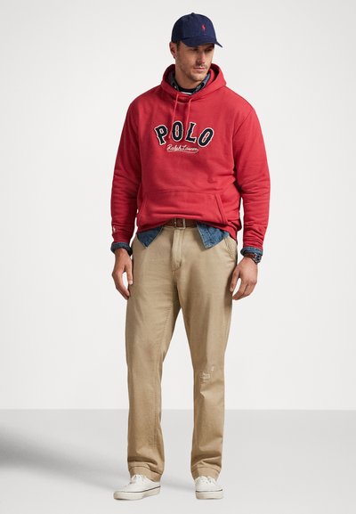 Άνδρας με κόκκινο φούτερ Polo, μπλε τζιν πουκάμισο, μπεζ παντελόνι, άσπρα αθλητικά παπούτσια, σκούφο ναυτικού μπλε και ρολόι, στέκεται μπροστά σε απλό φόντο.