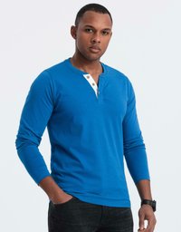 Man som bär en blå långärmad henley-tröja med vit knappslå och svarta jeans, står med ena handen i fickan.