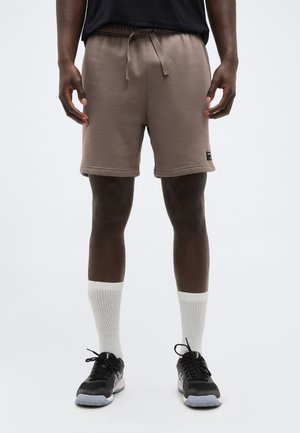 Személy barna zsinóros rövidnadrágot, fehér bokazoknit és fekete sportcipőt visel egyszínű világos háttér előtt.