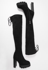 Paire de bottes plateformes noires en daim, montant au-dessus du genou, avec talons hauts épais, fermetures éclair latérales et détails à lacets sur le bord supérieur arrière.