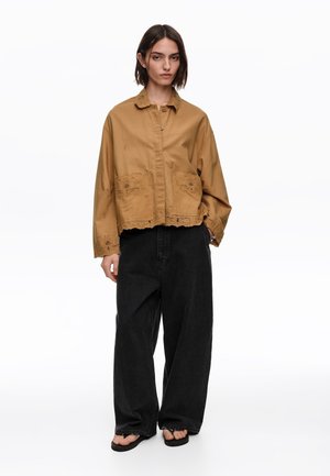 Kobieta w za dużej brązowej kurtce na guziki z haftem, luźnych czarnych spodniach i czarnych sandałach stojąca na białym tle.