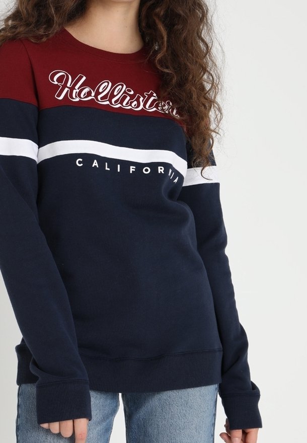 Personne portant un sweat Hollister California bleu marine, blanc et rouge à manches longues et un jean bleu clair.