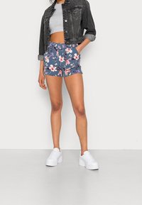 Blå shorts med blommönster, elastisk midja och fickor, i kombination med en grå crop top och en svart denimjacka, tillsammans med vita sneakers.