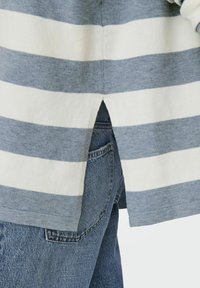 Maglione a righe blu e crema con dettaglio dell'orlo spaccato, abbinato a jeans in denim blu chiaro che mostrano la tasca posteriore. Tessuto morbido.