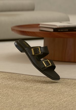 Sandale en cuir noir à enfiler avec deux boucles dorées, posée dans une pièce moquettée beige près d'une table basse ronde en bois avec des livres empilés.