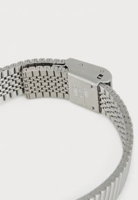 Montre en acier inoxydable avec un bracelet en maille tissée. Caractérisée par un fermoir poli avec des détails gravés. La finition globale est lisse et métallique.