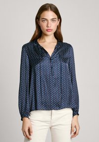 Ναυτικό σατέν μπλούζα με μακριά μανίκια, λαιμόκοψη σε V, κλείσιμο με κουμπί και λευκό και κόκκινο σχέδιο με πουά. Ελαστικές μανσέτες και χαλαρός στυλ.