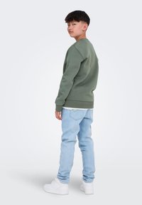 Sudadera de algodón verde con puños y dobladillo acanalados, combinada con jeans de mezclilla azul claro que presentan detalles en los bolsillos traseros y zapatillas blancas.