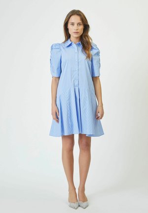 Himmelblaues Hemdkleid mit vertikalen weißen Streifen, gepufften Ärmeln, Knopfleiste und schwingendem Rock, aus einem glatten Stoff gefertigt. Silberne Akzent-Absätze.