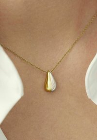 Collar de cadena de oro con un colgante en forma de lágrima en dos tonos, mitad de oro pulido y mitad de plata pulida, llevado sobre piel desnuda, encima de una prenda blanca.