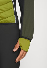 En tætsiddende jakke med en kviltet grøn front, sorte ærmer og en tommelfingerhulsmanchet. Materialet ser lette ud og har en mat tekstur.