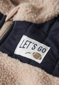 Fleece jack met marineblauwe en lichtroze materialen; heeft een "LET'S GO" patch en een bruine voetbalfoto. Zachte textuur met een ritssluiting.