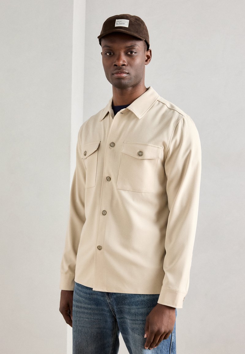 Μπεζ πουκάμισο με κουμπιά και δύο τσέπες στο στήθος, κατασκευασμένο από λείο ύφασμα. Συνδυασμένο με μπλε τζιν και καφέ καπέλο. Απλός σχεδιασμός, χωρίς μοτίβα.