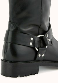 Botte de cheville en cuir noir avec une large sangle en métal argenté, finition texturée et semelle rugged.