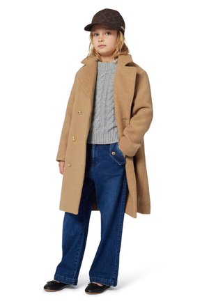 Ein Kind trägt einen braunen Mantel, einen grauen Strickpullover, weite Jeans, schwarze Schuhe und eine dunkelbraune gemusterte Mütze. Es steht mit einer Hand in der Tasche.