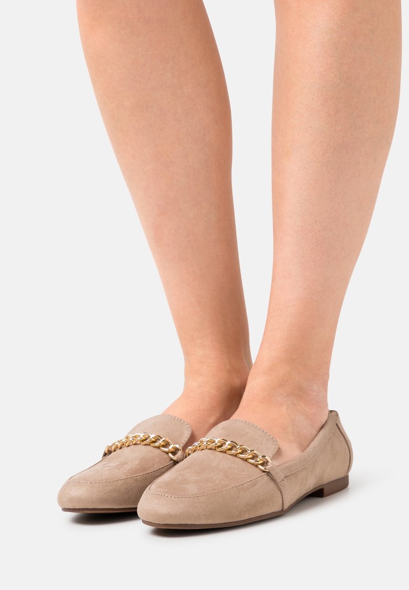Chaussures plates en daim beige avec détail de chaîne dorée, portées pieds nus, présentées sur un fond blanc.