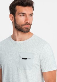 Světle šedé tričko s krátkým rukávem s jemným vzorem textu po celém povrchu, s kontrastní kapsou a černým akcentem.