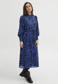 Blau-schwarz gemustertes Kleid mit langen Ärmeln, Kragen, Knopfleiste und tailliertem Schnitt; kombiniert mit schwarzen Knöchelstiefeln.