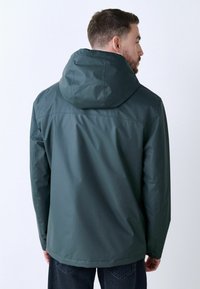 Donker groene capuchonjack gemaakt van glad, waterafstotend materiaal. Beschikt over een eenvoudig ontwerp zonder zichtbare patronen of hardware.
