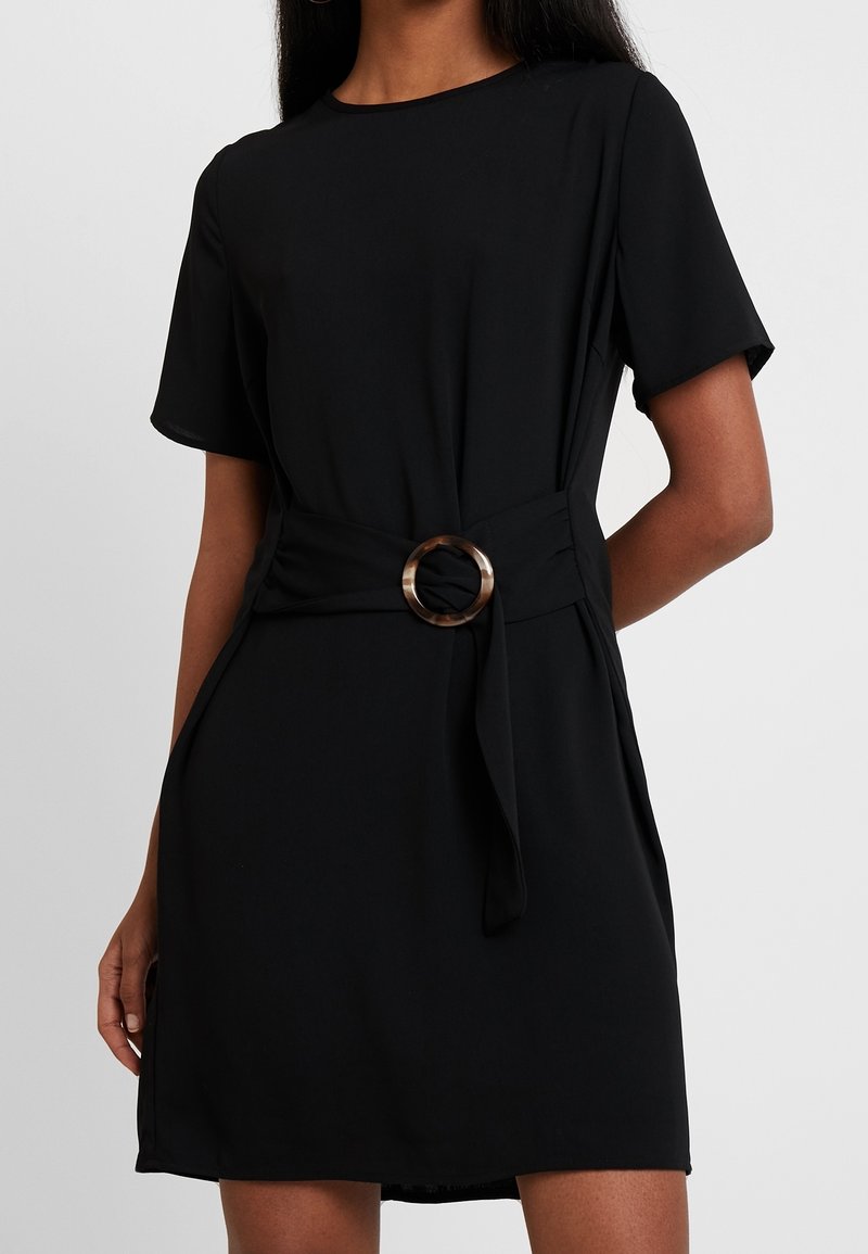 Femme portant une robe noire à manches courtes avec une boucle de ceinture ronde en écaille de tortue à la taille, les mains reposant le long du corps.