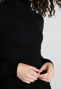 Maglione dolcevita nero con texture a costine e design aderente, dotato di maniche lunghe e polsini attillati. Mani incrociate all'altezza della vita.