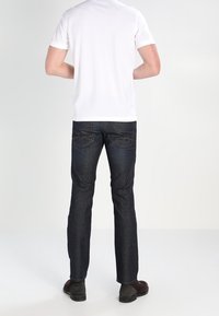 T-shirt blanc à manches courtes associé à un jean bleu foncé. Le jean possède des poches arrière avec une couture distinctive. Les chaussures sont brunes avec une texture lisse.