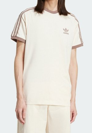 Person trägt ein cremefarbenes Adidas T-Shirt mit braunen Streifen an den Ärmeln und Logo auf der Brust, kombiniert mit passenden cremefarbenen Shorts.