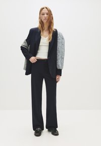 Jonge vrouw met lang rood haar, gekleed in een marineblauw streepjespak, een witte cropped top, een grijze trui nonchalant om haar schouders en zwarte schoenen.