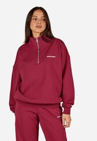 Burgundijas krāsas rāvējslēdzēja sporta džemperis ar augstu apkakli, elastīgām aprocēm un balto logo "PEGADOR" kreisajā krūtī. Mīksta, teksturēta auduma.