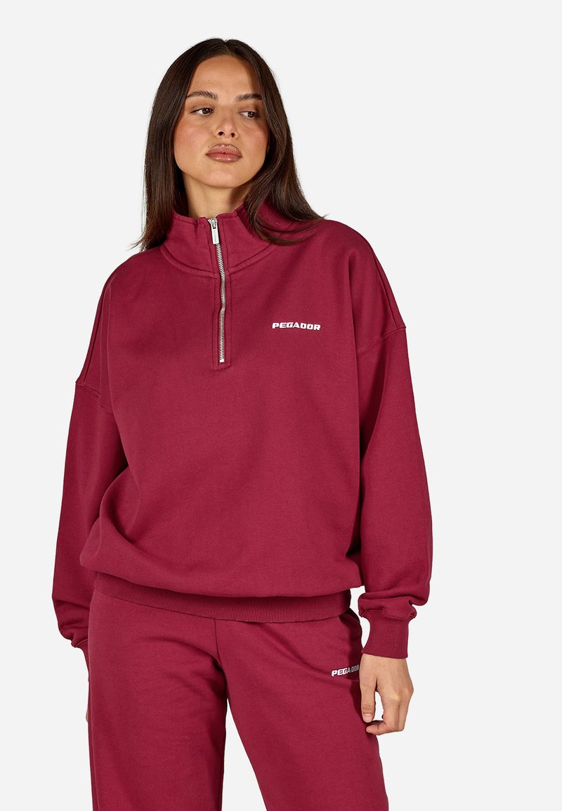 Burgundijas krāsas rāvējslēdzēja sporta džemperis ar augstu apkakli, elastīgām aprocēm un balto logo "PEGADOR" kreisajā krūtī. Mīksta, teksturēta auduma.