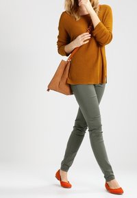 Sinappikeltainen neulepusero, oliivinvihreät joustavat housut, oranssit balleriinat ja tan-värinen olkalaukku hihnalla. Malli seisoo neutraalilla taustalla.