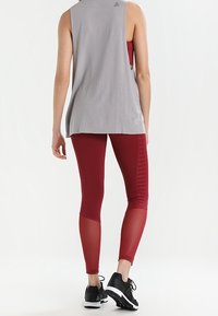 Top grigio senza maniche abbinato a leggings rossi con un design testurizzato e dettagli in rete, completato da scarpe sportive nere.
