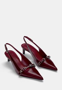 Talons hauts slingback en cuir verni bourgogne avec des pointes effilées, un détail central en chaîne métallique et des talons larges de 5 cm, présentant une texture lisse.