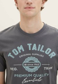 Graues Baumwoll-T-Shirt mit auffälliger blauer Typografie und einem kreisförmigen Logo mit Text, runder Ausschnitt und kurzen Ärmeln.
