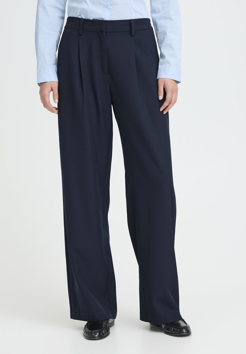 Pantalon large marine en tissu lisse, doté d'une taille haute, de plis à l'avant et de passants pour ceinture, associé à des ballerines noires.