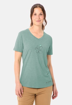 Frau trägt ein grünes V-Ausschnitt-T-Shirt mit floralem Muster und beigefarbenen Hosen, steht mit neutralem Gesichtsausdruck vor einem schlichten Hintergrund.