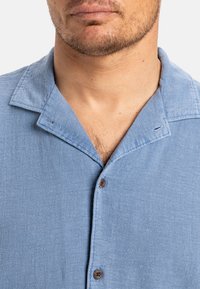 Svetlo modrá košeľa s krátkym rukávom vyrobená z textúrovanej tkaniny, s golierom, gombíkmi na prednej strane a dvoma hnedými gombíkmi na zapínanie.