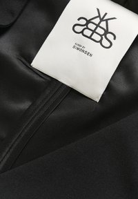 Sort stof med en glat tekstur, prydet med et hvidt label med "KAREN BY SIMONSEN" og et grafisk logo, fastgjort nær sømmen.