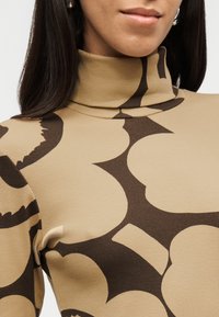 Γυναίκα που φοράει μπεζ ζιβάγκο με μεγάλα σκούρα καφέ αφηρημένα λουλουδένια σχέδια, εμφανιζόμενη από τους ώμους έως το στόμα.