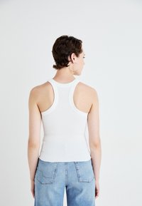 Débardeur blanc côtelé avec un design dos de nageur, présentant une silhouette ajustée. Associé à un denim bleu clair, avec une coupe détendue et des poches visibles.