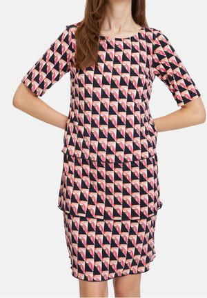 Kobieta ubrana w dopasowaną bluzkę i spódnicę z geometrycznym wzorem w kolorach czarnym, różowym i białym, stojąca z rękami na biodrach.