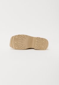 Bruine rubberen schoenzool met zigzagpatroon en afgeronde randen, met de merknaam "UGG" in het midden geperst.
